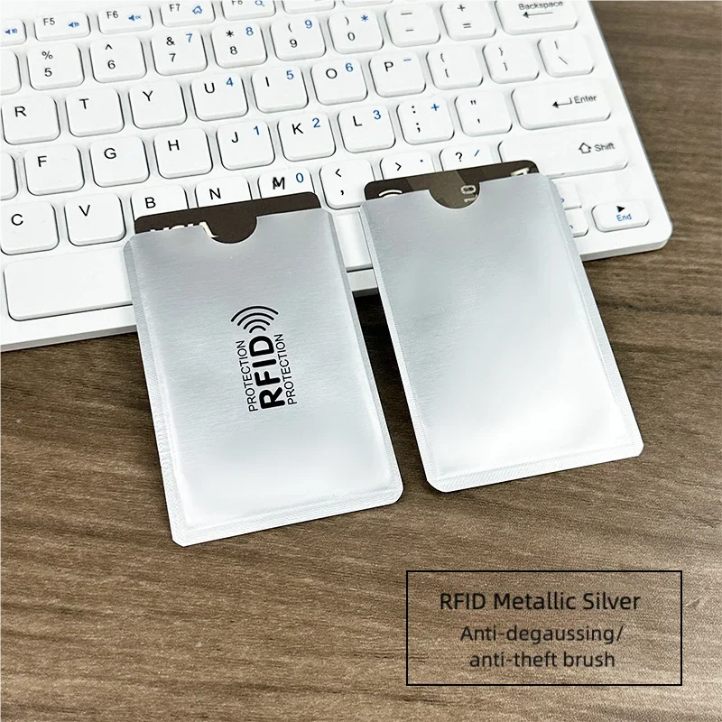 Two metallic silver RFID blocking card sleeves placed on a wooden surface next to a laptop keyboard.