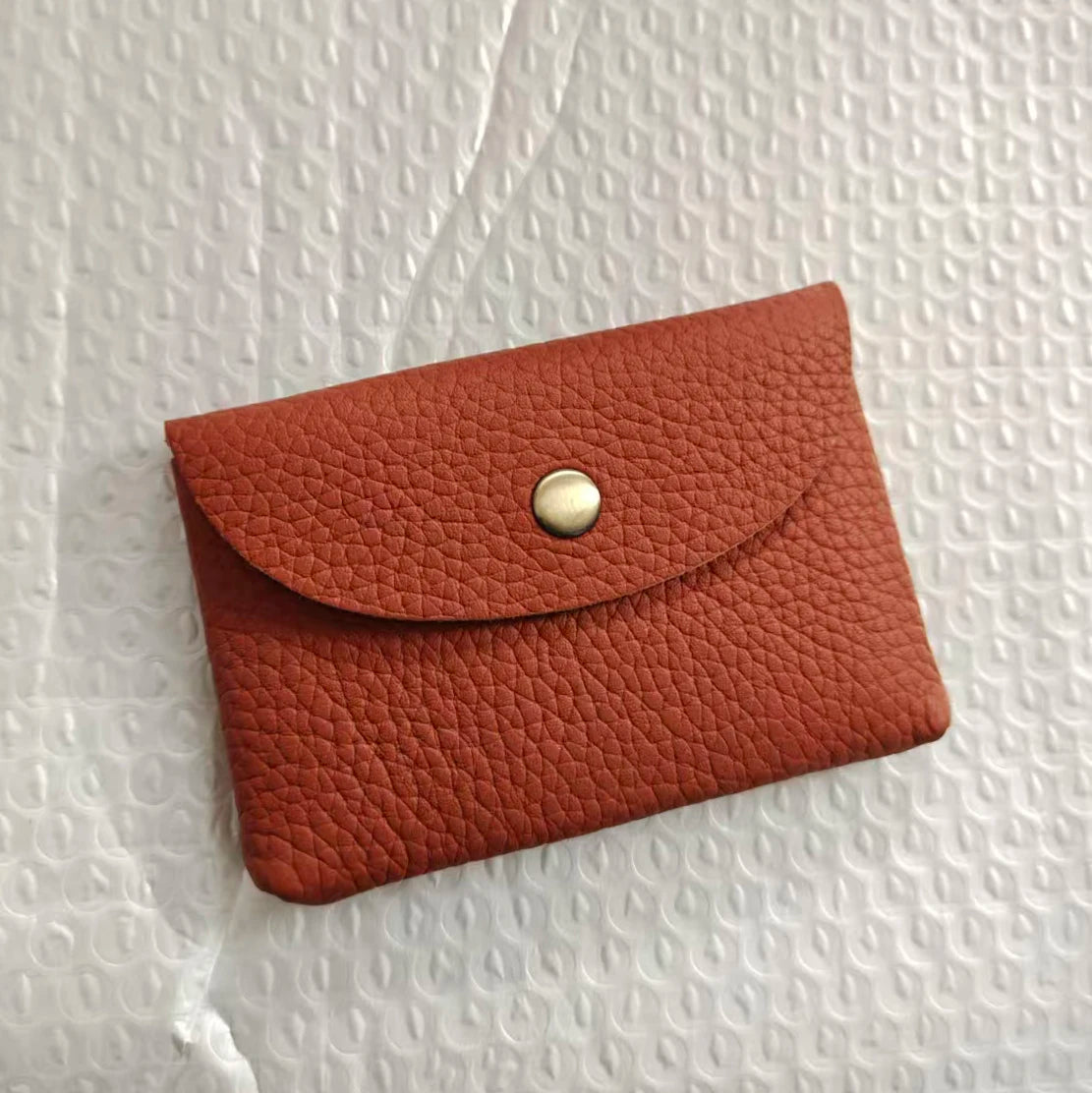 A terracotta-colored genuine leather card holder with a gold-toned clasp resting on a white textured surface.