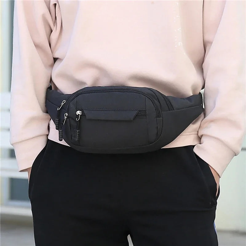 A black canvas waist fanny bag is held by a person wearing a light pink top and black pants.