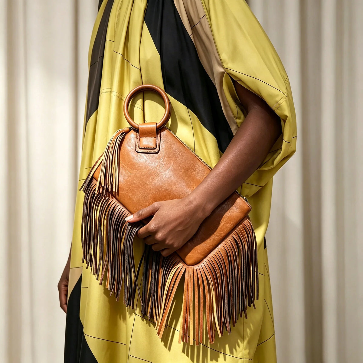 A woman holds a brown fringed clutch bag with tassels against a yellow and black striped garment.