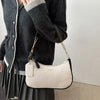 A light beige leather crescent crossbody bag with a gold chain strap is shown being held by a person wearing a dark sweater.
