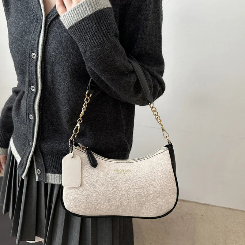 A light beige leather crescent crossbody bag with a gold chain strap is shown being held by a person wearing a dark sweater.