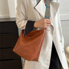 A person holds a tan leather shoulder bag with an embroidered blue and orange strap under a white blazer.