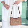 A woman holds a brown leather crossbody bag with a gold chain strap and a diamond pendant against a white dress.