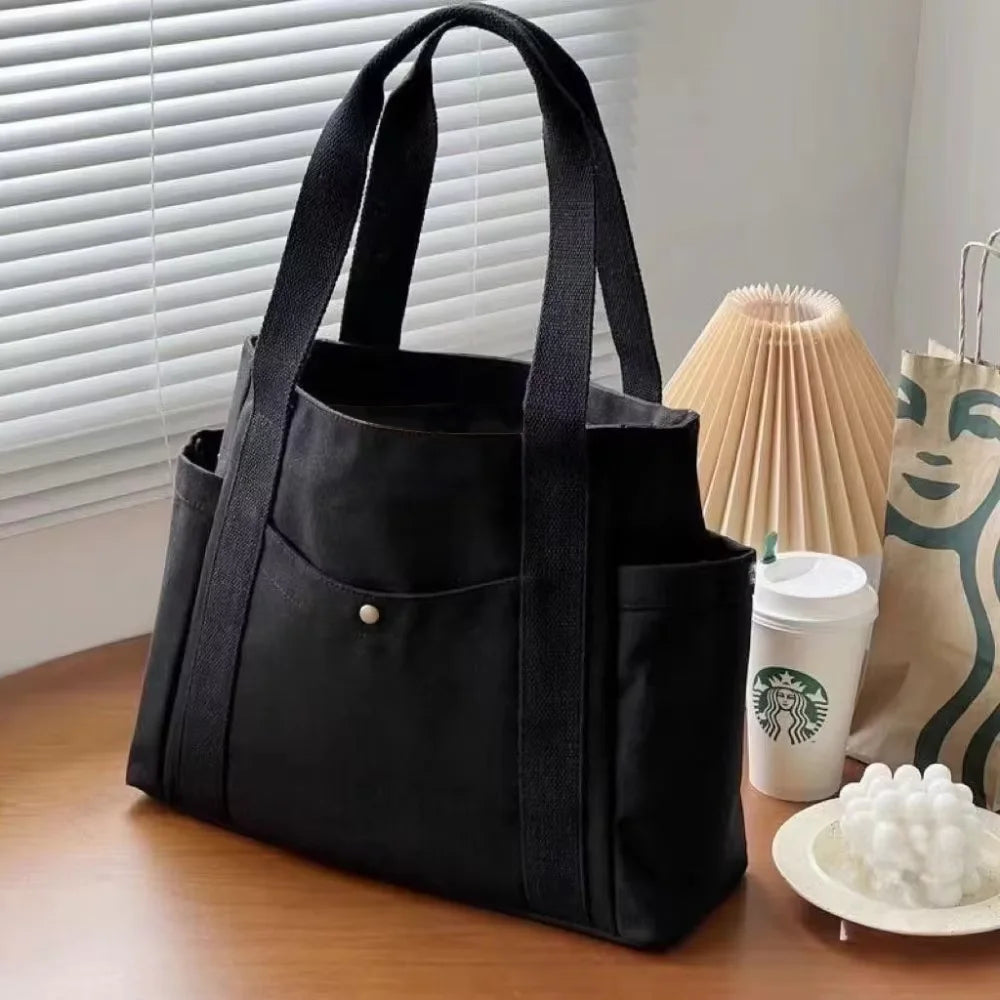 Bolso tote de lona negra con tirantes apoyado sobre una superficie de madera junto a una planta en maceta.