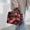 A person holds a black floral patterned cloth tote bag with pink and burgundy roses against a light background.