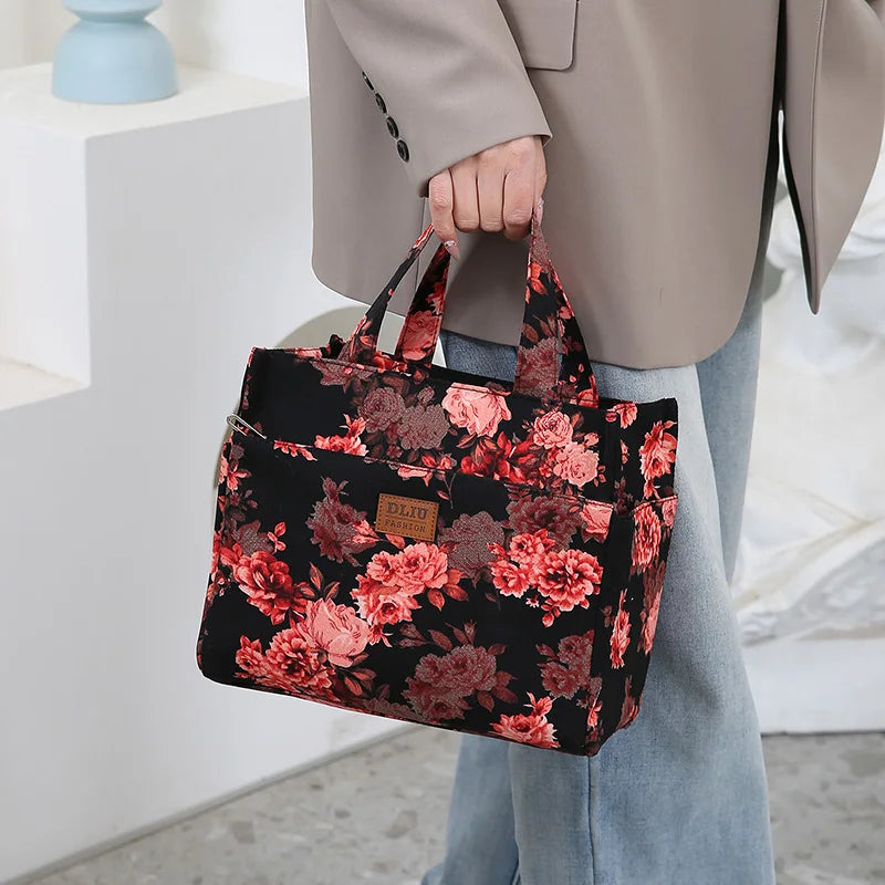 A person holds a black floral patterned cloth tote bag with pink and burgundy roses against a light background.