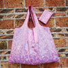 A pink reusable shopping bag is displayed against a brick wall.