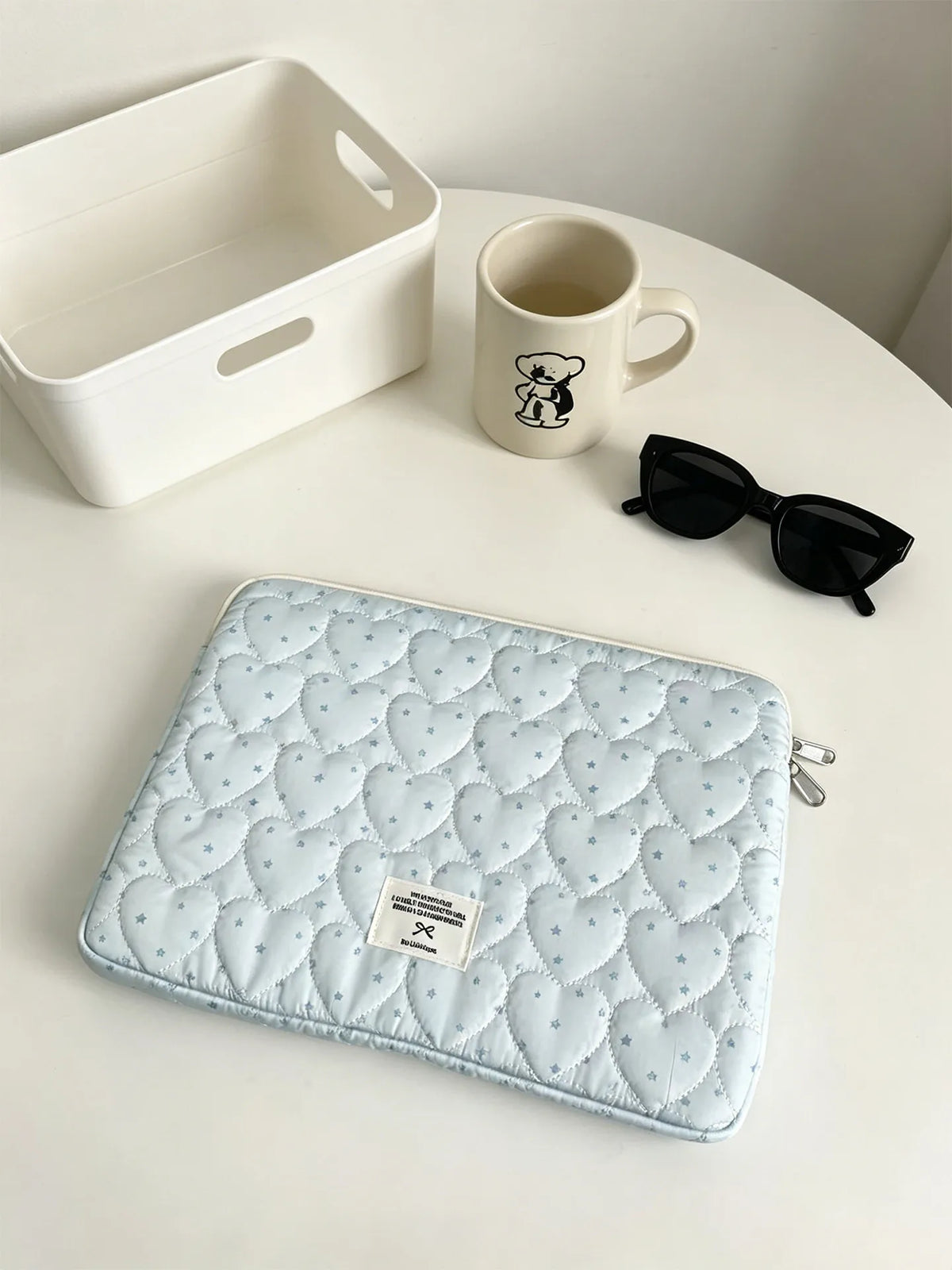 A light blue quilted laptop sleeve with a pattern of small hearts is displayed on a white table next to a mug and sunglasses.