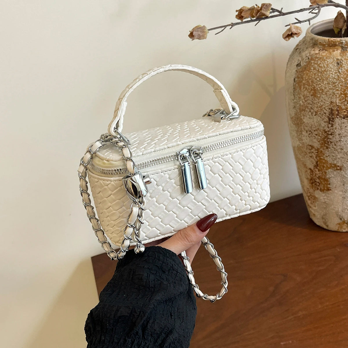 A white woven crossbody bag with silver chain straps is held by a hand against a light background.