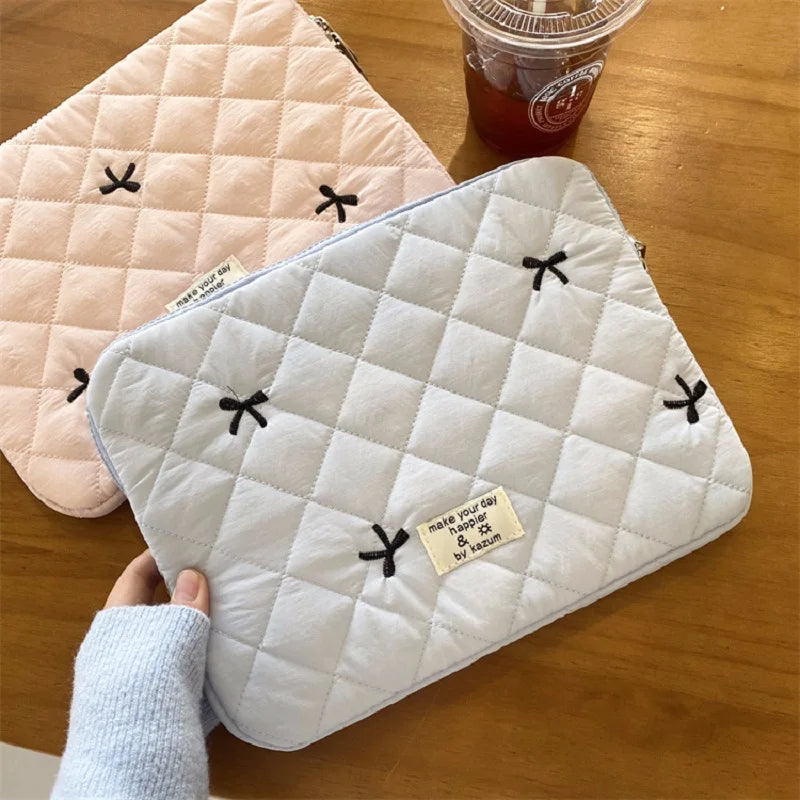 A light blue quilted laptop sleeve with black tie-shaped embroidery is shown next to a pink quilted fabric and a glass jar.