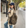 A young woman wearing a beige jacket and jeans walks down a tree-lined street carrying a leopard print canvas backpack.