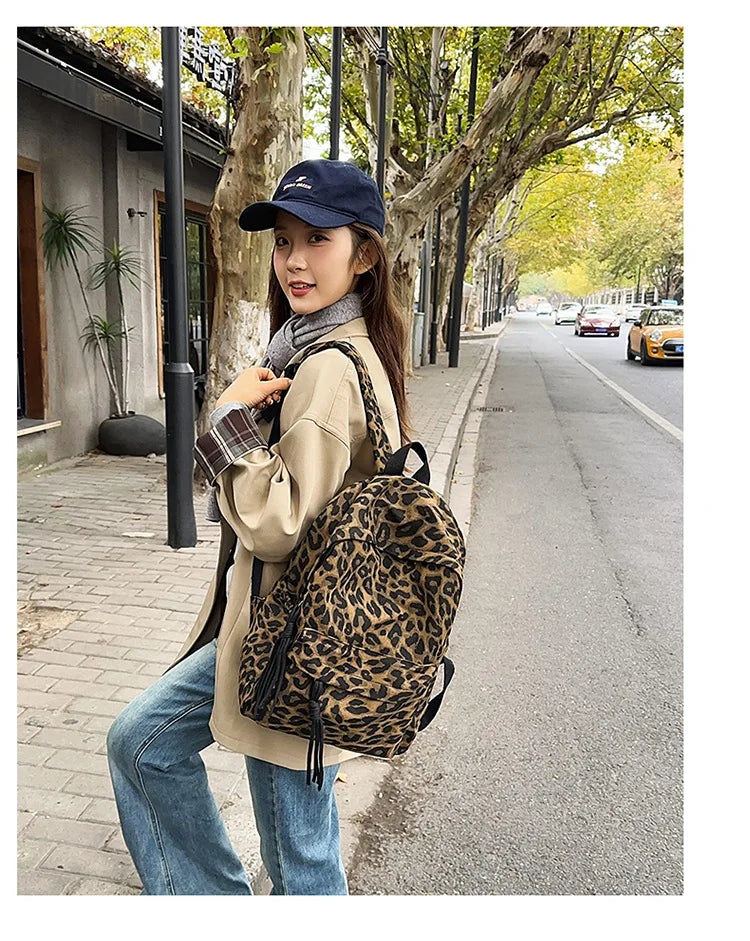 A young woman wearing a beige jacket and jeans walks down a tree-lined street carrying a leopard print canvas backpack.