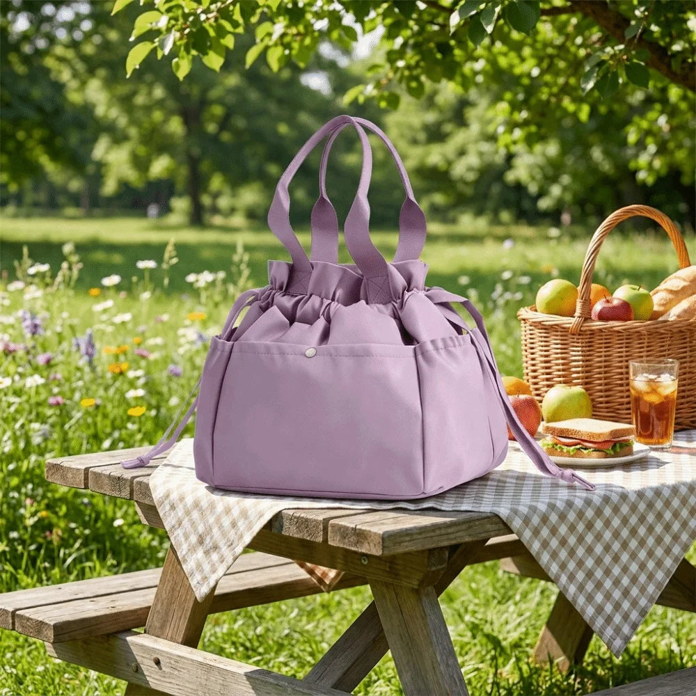 A purple insulated drawstring lunch bag with multi-pocket storage resting on a wooden picnic table in a sunny outdoor setting