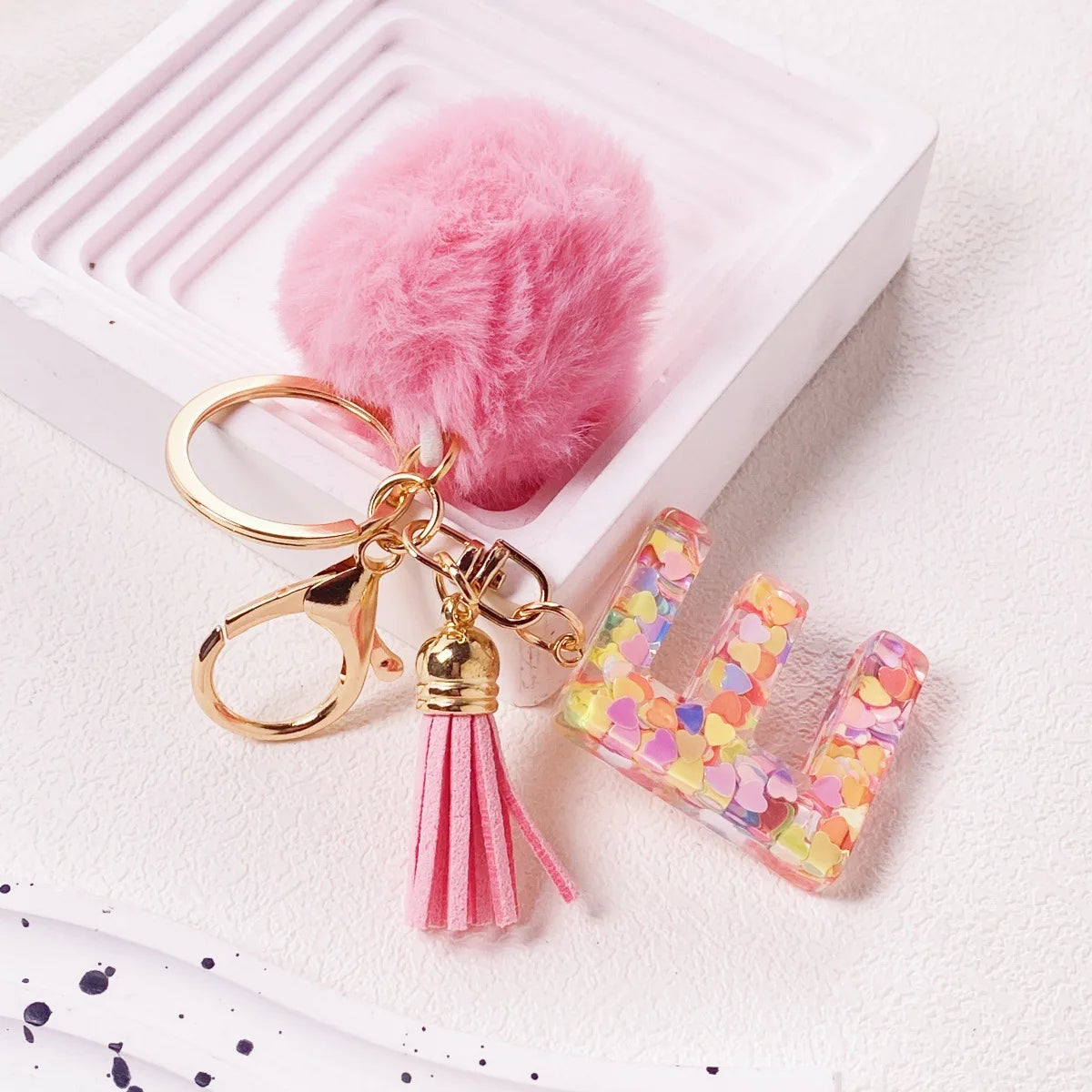 A shiny resin initial letter keychain with a pink alphabet charm and a fluffy pink pom-pom resting in a white tray.