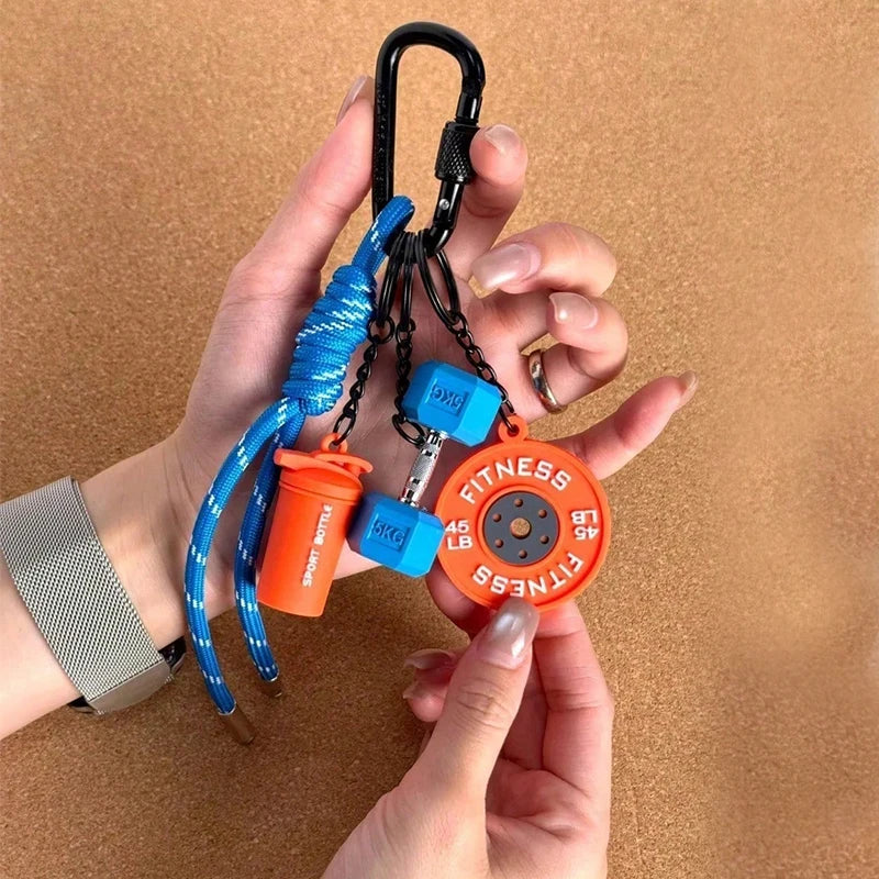 A hand holds a black nylon fitness keychain with an orange fitness disc and blue rope for gym or outdoor gear.