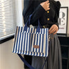 Une femme tient un sac bandoulière en toile rayée bleu marine et blanc devant une fenêtre avec des stores horizontaux.