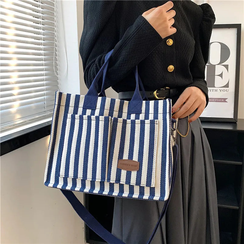 Une femme tient un sac bandoulière en toile rayée bleu marine et blanc devant une fenêtre avec des stores horizontaux.