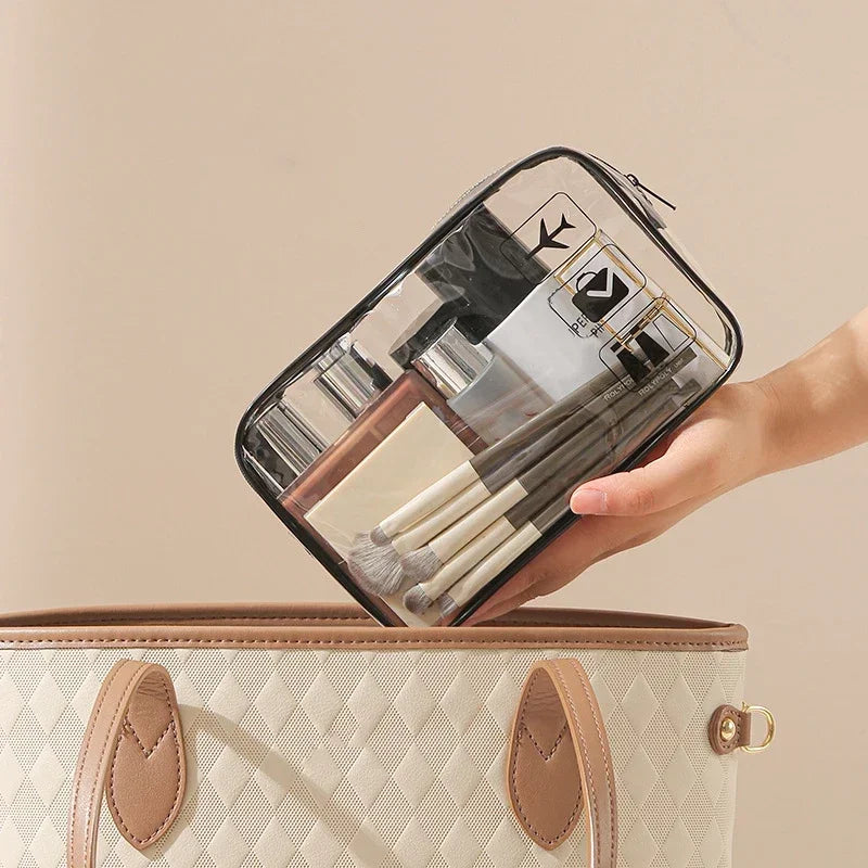 A hand holds a clear PVC toiletry bag filled with makeup brushes and other toiletries above a beige quilted handbag.