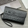 A black crocodile pattern clutch bag resting on a gray wooden surface next to a silver laptop and white smartphone.