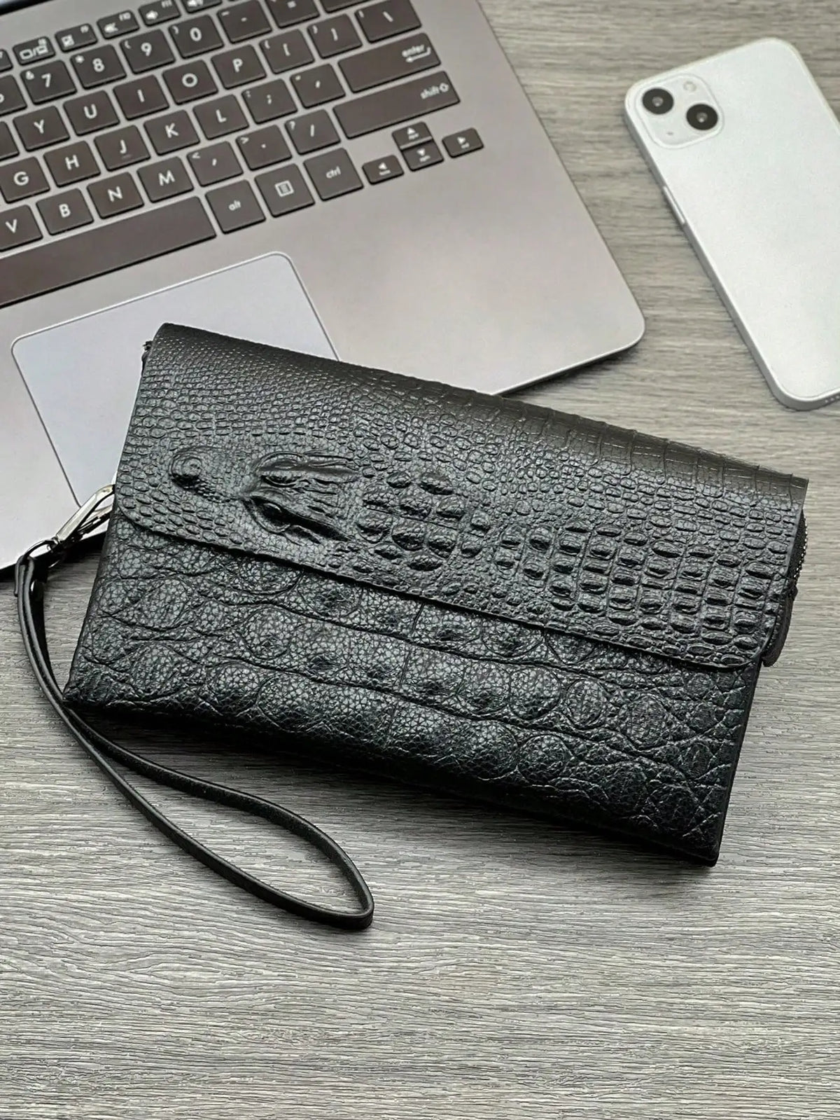A black crocodile pattern clutch bag resting on a gray wooden surface next to a silver laptop and white smartphone.
