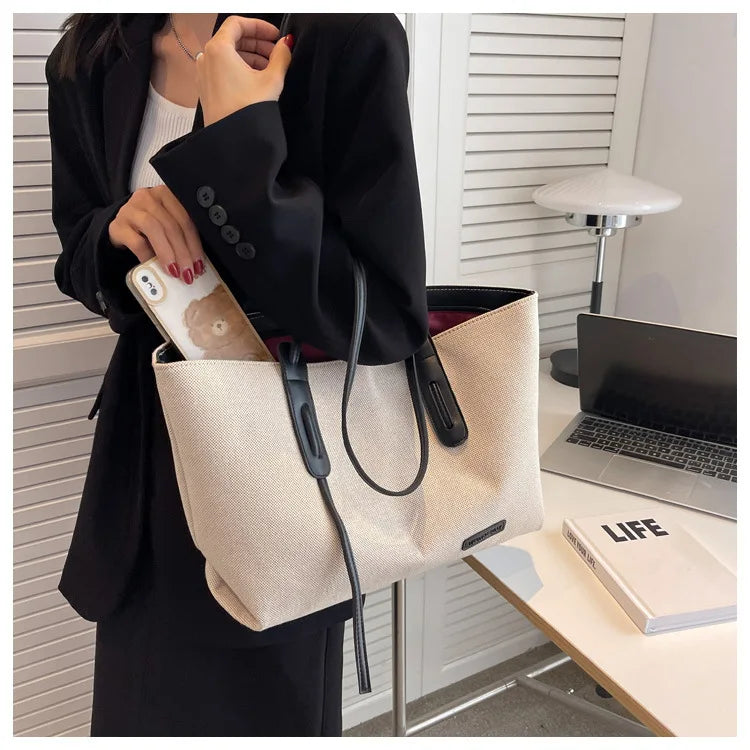 A woman holds a large beige canvas shoulder tote bag with a black strap and a smartphone in a modern indoor setting.