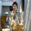 A woman models a tan cotton canvas shoulder crossbody bag with plaid shirt and white pants outdoors.