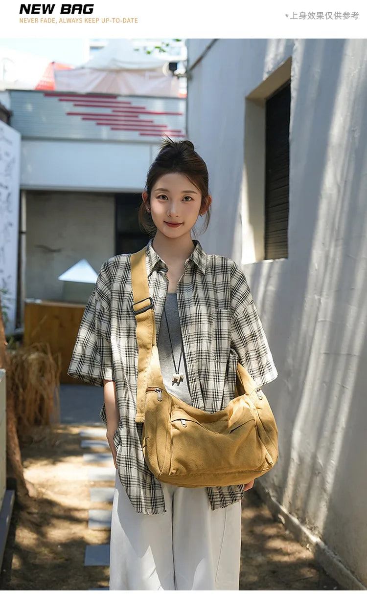 A woman models a tan cotton canvas shoulder crossbody bag with plaid shirt and white pants outdoors.