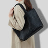 A woman holds a black woven vegan leather tote bag with a textured pattern against a light beige coat.