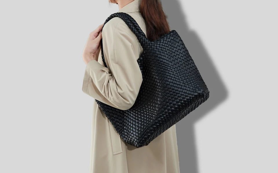A woman holds a black woven vegan leather tote bag with a textured pattern against a light beige coat.