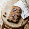 A brown leather zipper wallet featuring a Highland cow print design resting on a wooden surface next to some dried branches a