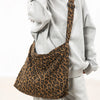 A woman is holding a brown leopard print canvas shoulder bag with an adjustable strap against a white background.