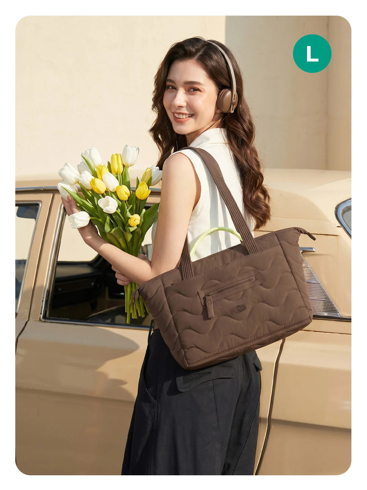A woman holds a brown quilted tote bag with a detachable strap and a bouquet of white and yellow tulips next to a vintage car