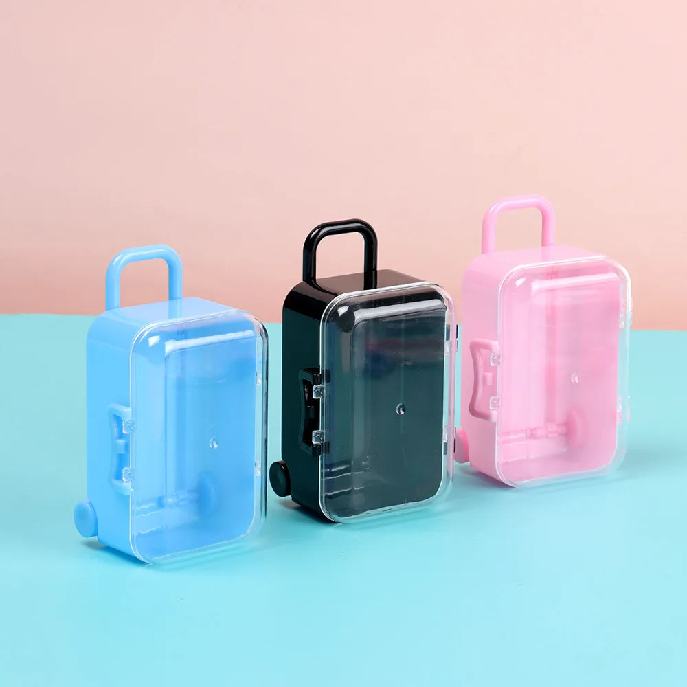 Three miniature doll suitcases in blue, black, and pink plastic are displayed on a light blue surface.