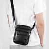 A black leather messenger bag with a slanted back worn by a man in a white t-shirt.