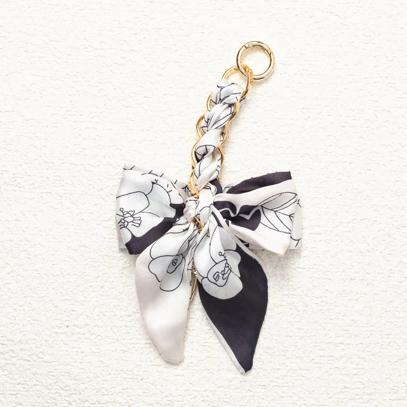 A white and black floral patterned fabric bow attached to a gold chain strap extension for a handbag.