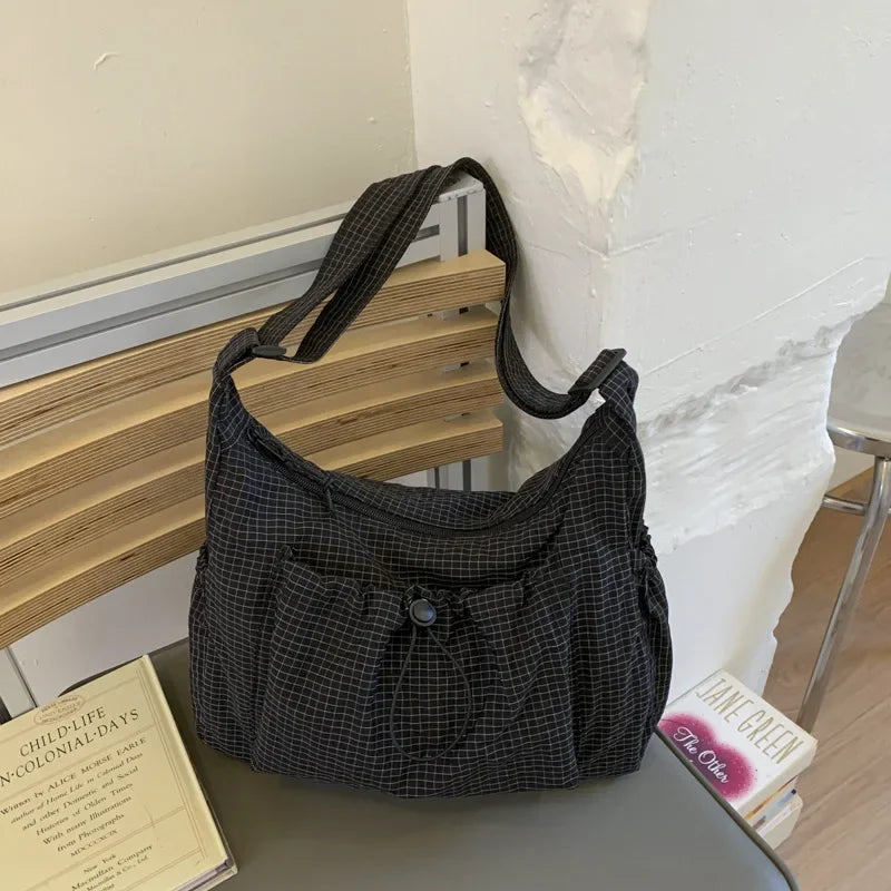 A dark plaid nylon drawstring tote bag resting on a wooden shelf next to a white wall.