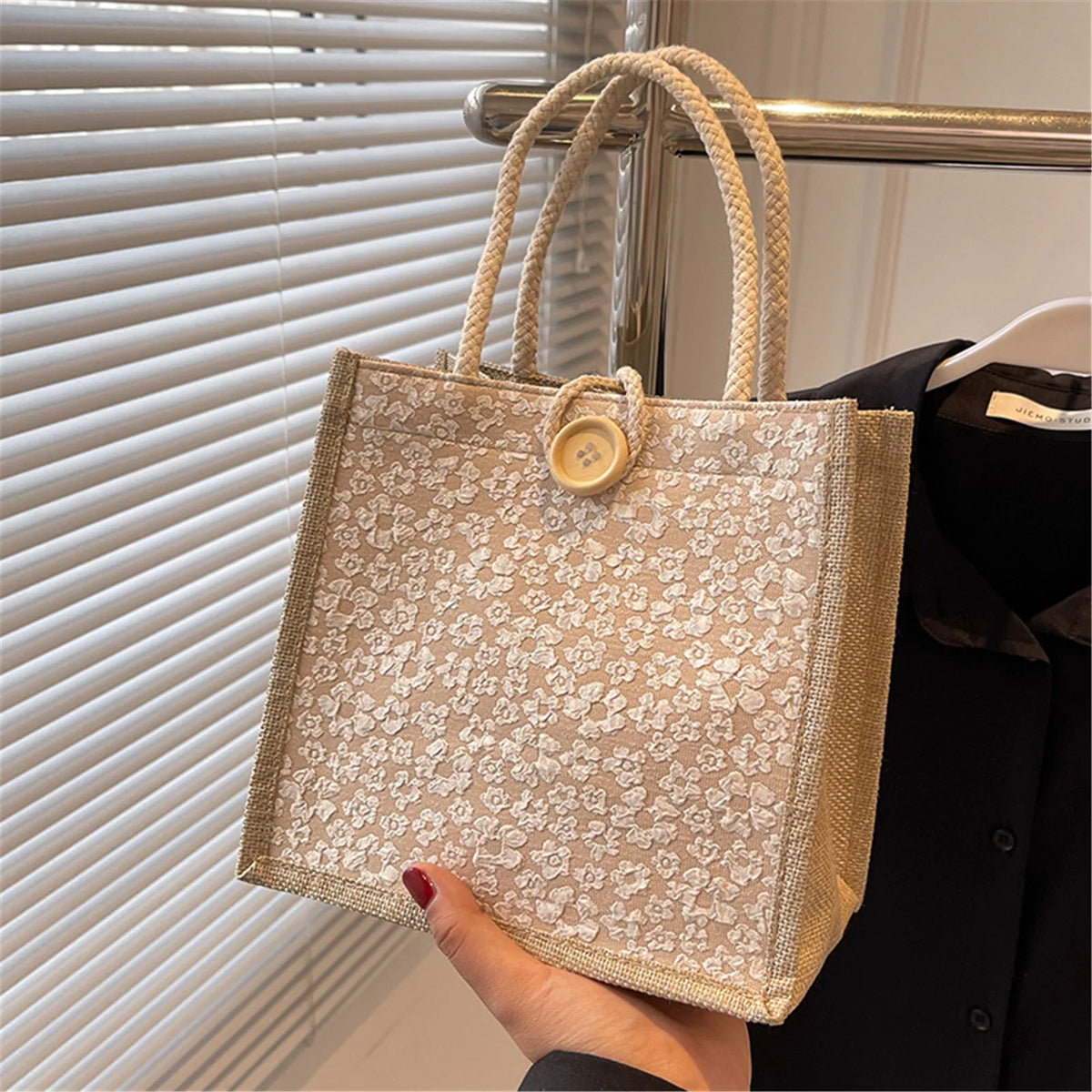 A beige floral linen tote bag with woven handles and a wooden closure is displayed against a window blind.