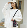 A woman wearing a white sweatshirt and light wash jeans holds a black nylon crossbody phone bag.
