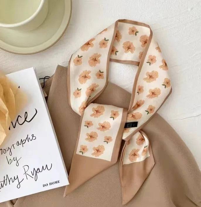 Foulard de cheveux floral en soie beige drapé sur un tissu marron, à côté d'un carnet.