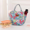 Una bolsa térmica de bento estampada y colorida con asa, junto a un pequeño recipiente de comida naranja y una botella de col