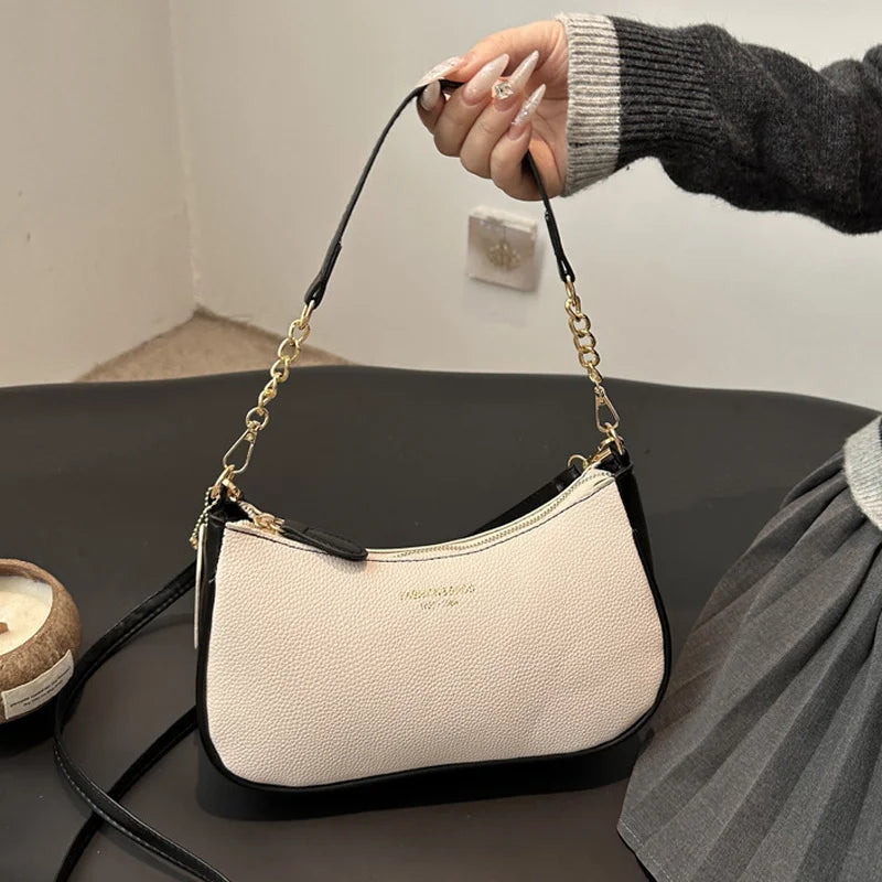 A close-up of a cream and black crescent crossbody bag with gold hardware being held by a hand wearing a gray sweater.