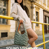 A woman wearing a beige hoodie holds a green quilted nylon shoulder bag outdoors.