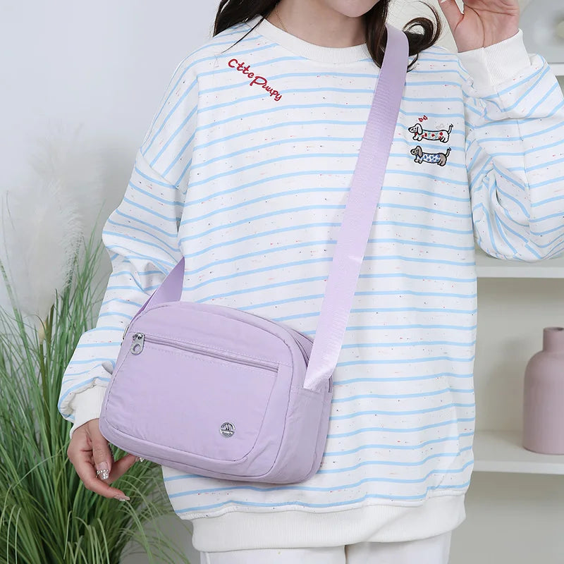Eine Frau hält eine helllila Nylon-Crossbody-Tasche vor einem weißen gestreiften Sweatshirt.
