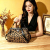 A woman holds a leopard print faux leather handbag with a top handle and crossbody strap on a wooden surface.
