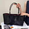 A black crocodile embossed leather tote bag with a magnetic closure being held by a person.