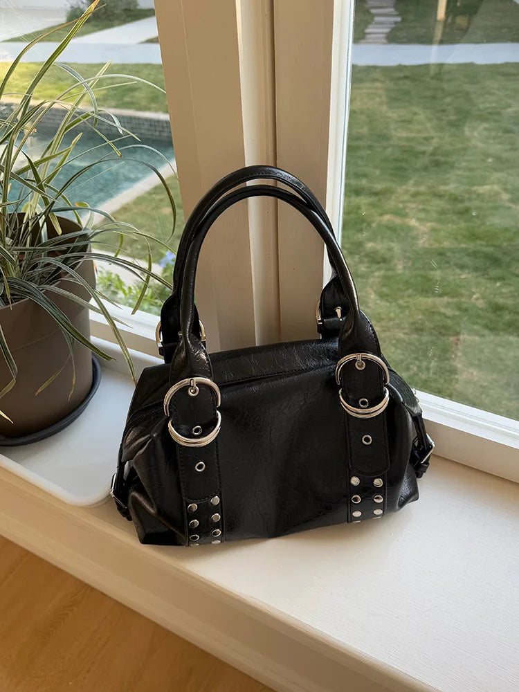 A black PU leather shoulder bag with silver rivets and hardware resting on a windowsill next to a potted plant.
