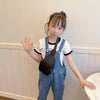 A young girl with dark hair is holding up her hand in a wave while wearing a black nylon crossbody bag over a white t-shirt a