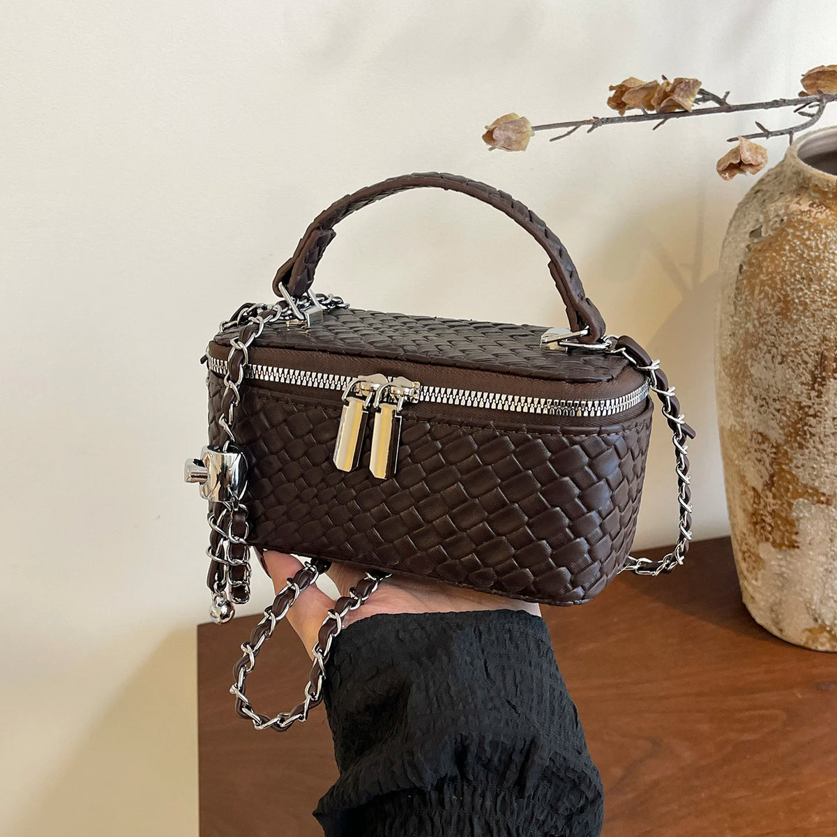 A dark brown woven crossbody bag with a silver chain strap is held against a light background.