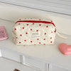 A white quilted makeup bag with red piping and a pink heart design resting on a white shelf.